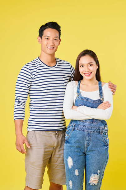 Positive young Vietnamese man is striped shirt hugging his pretty girlfriend wearing denim overall - 写真・画像
