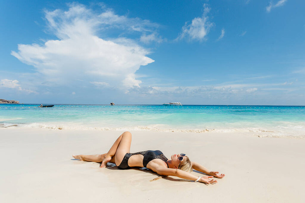 Blonde young woman in black swimsuit lying on side at beach. Beautiful tanned girl lying on sand. Attractive girl wearing glamour swimwear on tropical beach while sunbathing. - Photo, Image