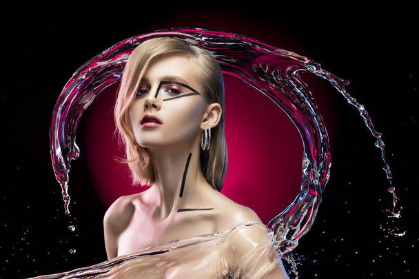 Beautiful blonde girl with naked shoulders and avant-garde conceptual makeup, surrounded by splashes and drops of water on a black and red background. Copy space. Advertising and commercial design. - Photo, image