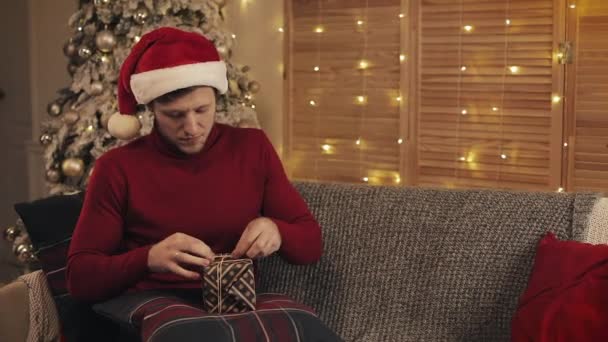 Close Up of Young Handsome Man Wearing a Santas Hat sitting on a Sofa near Christmas Tree at Living Room Ties a Bow on a Present Box than Looks to Camera and Smiles. Concept of Holidays and New Year. - Video