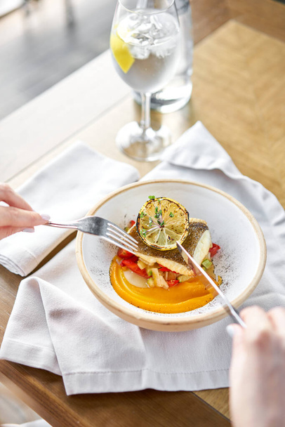 Roasted Halibut with vegetables, paprika pepper and pumpkin cream. Lunch in a restaurant, a woman eats delicious and healthy food. Dish decorated with a slice of lemon. Restaurant menu - Φωτογραφία, εικόνα