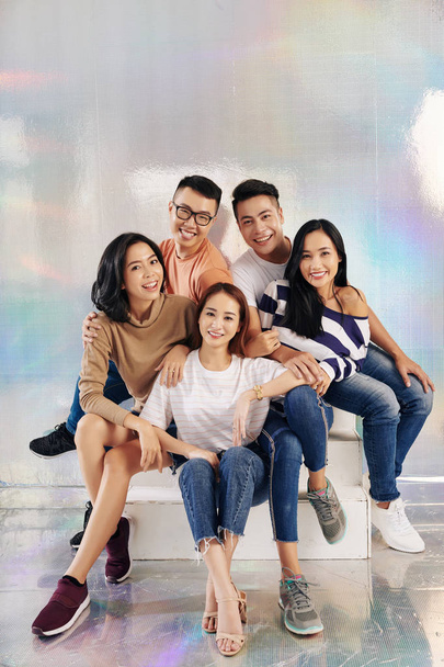 fröhliche junge asiatische Menschen in lässiger Kleidung posieren gemeinsam auf Stufen im Studio mit holographischem Hintergrund - Foto, Bild