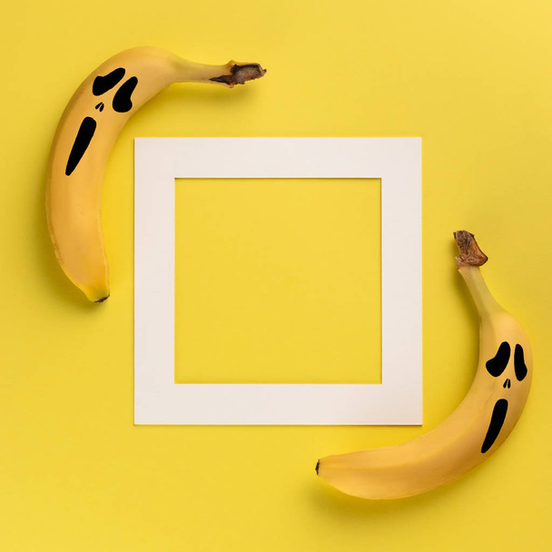 Halloween concept with two bananas with painted on them scary mask of fear and white frame with copy space for text on yellow background - Foto, imagen