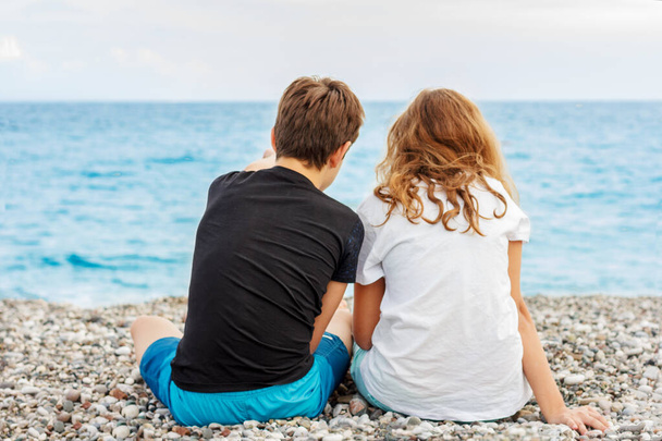 Un paio di bei ragazzi, il primo amore. Guy abbraccia una ragazza seduta sulla spiaggia di ciottoli uno accanto all'altro e guarda il mare. Spara da dietro
 - Foto, immagini