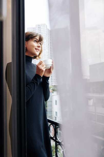 Pensive young woman drinking cup of coffee and enjoying the view from her balcony - 写真・画像