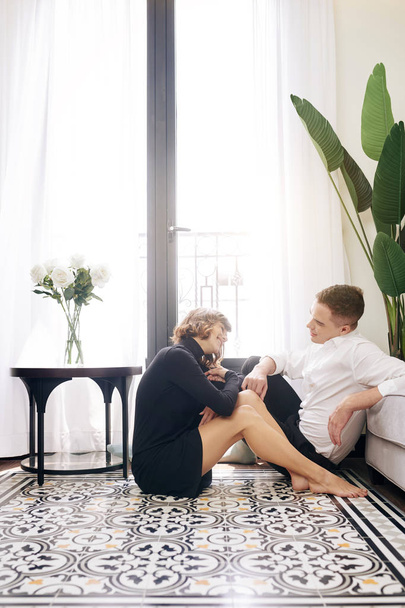 Positivo jovem casal apaixonado descansando no chão na sala de estar e conversando
 - Foto, Imagem