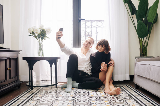 Joyeux jeune homme assis sur le sol à la maison avec sa petite amie et prendre selfie
 - Photo, image