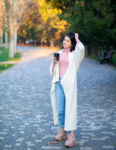 Sonbahar parkında beyaz hırkalı genç bir kadın, elinde siyah bir fincan kahveyle. - Fotoğraf, Görsel