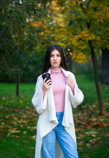 young female in white cardigan in the autumn park with black cup of coffee - Foto, imagen