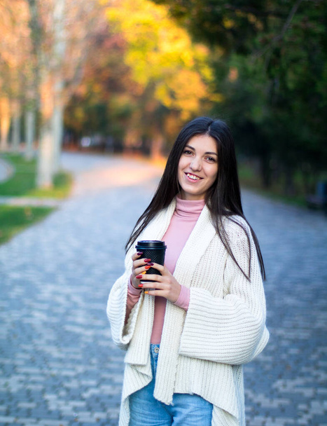 young female in white cardigan in the autumn park with black cup of coffee - Foto, Bild