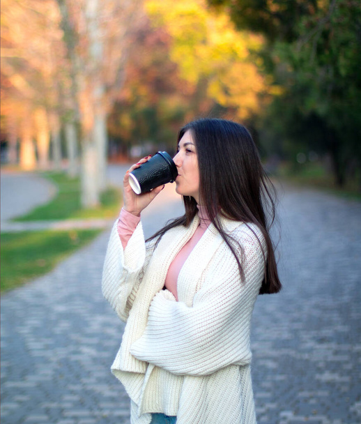 hembra joven en cárdigan blanco en el parque de otoño con taza de café negro
 - Foto, Imagen