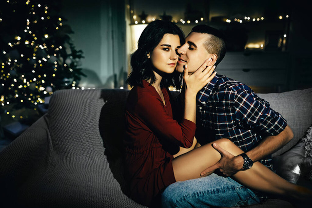 happy, couple in love in the dark, evening on the couch, dark background, christmas tree lights. Christmas evening. New Year. hug and kiss. attraction,romantic evening and date. - Fotoğraf, Görsel