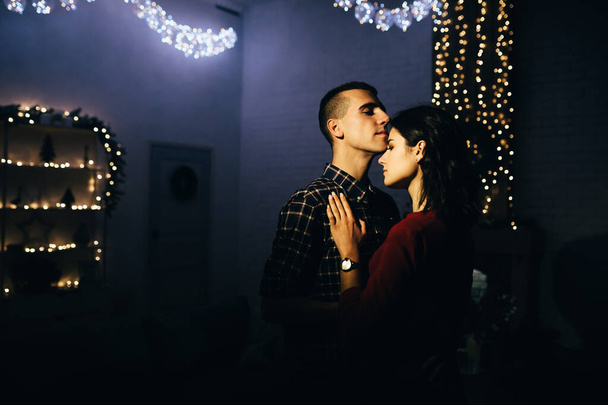 Ευτυχισμένο ζευγάρι ερωτευμένο στο σκοτάδι, βράδυ στον καναπέ, σκοτεινό φόντο, φωτάκια χριστουγεννιάτικου δέντρου. Χριστουγεννιάτικο βράδυ. Πρωτοχρονιά. Αγκαλιά και φιλί. έλξη, ρομαντικό βράδυ και ημερομηνία. - Φωτογραφία, εικόνα