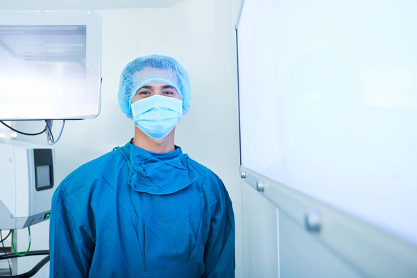 Retrato de jovem cirurgião estagiário vietnamita positivo em esfregaços, máscara protetora e boné em pé na sala de cirurgia
 - Foto, Imagem
