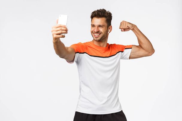 Kestävyyttä, urheilua, terveellisiä elämäntapoja. Komea nuori mies tyytyväinen omaan kehon muotoon, osoittaa lihas ottaa selfie, pidä älypuhelin hymyilee tyytyväinen, käyttää langattomia nappikuulokkeet valkoinen tausta
 - Valokuva, kuva
