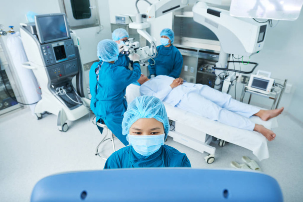 Вьетнамский хирург-ассистент с помощью компьютера манипулирует оборудованием над пациентом на кровати и готовится к операции
 - Фото, изображение
