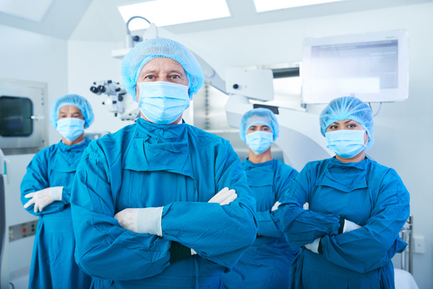 Equipe confiante de cirurgiões em esfregaços, bonés e máscaras faciais de pé com os braços dobrados e olhando para a câmera
 - Foto, Imagem