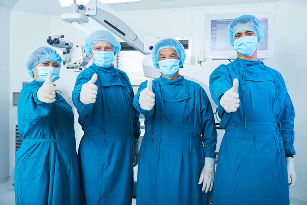 Equipo de cirujanos en matorrales y máscaras faciales que muestran pulgares hacia arriba después de una cirugía incesante
 - Foto, imagen
