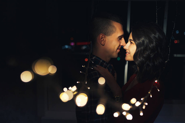 paar verliefd in het donker tussen de lichten van bloemenslingers knuffels en kussen. vakantie en datum voor Kerstmis. Twaalf uur, kus op oudejaarsavond. in de lichten op de achtergrond van het raam. gelukken - Foto, afbeelding