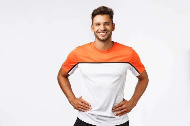 Fitness, sports and active lifestyle concept. Studio portrait handsome young male trainer in activewear, hold arms on waist, smiling friendly and determined, get your head in game, ready to workout - Photo, Image