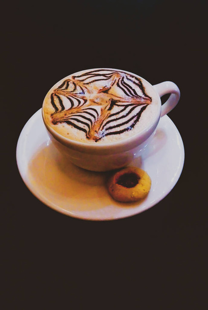 Taza de café capuchino con diseo de chocolate en forma de asterisco y galleta glaseada, sobre fondo oscuro
. - Foto, Imagen
