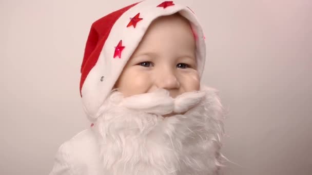 funny three-year-old caucasian kid with artificial beard in Santa Claus hat with blinking red lights smiling on white background. father straightenes his mustache. costume for celebrating christmas - Filmagem, Vídeo