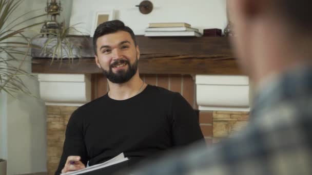 Close-up face of an adult Caucasian man sitting with papers, talking, and smiling. Professional male psychologist telling story to patient sitting opposite. Camera moving from right to left. - Filmagem, Vídeo