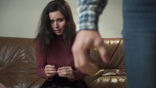 Portrait of a frightened to death Caucasian woman sitting at the couch with ring and hiding her face behind hands. Man at the foreground compressing his hand into fist. Domestic violence, tyranny. - Materiaali, video