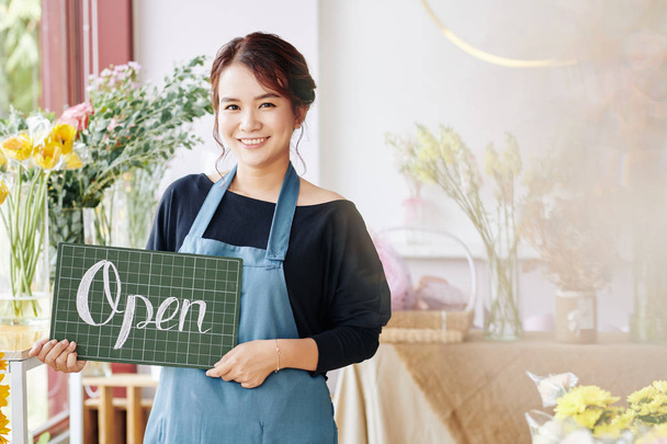 Pretty young female flower shop owner holding open sign and smiling at camera - Photo, Image