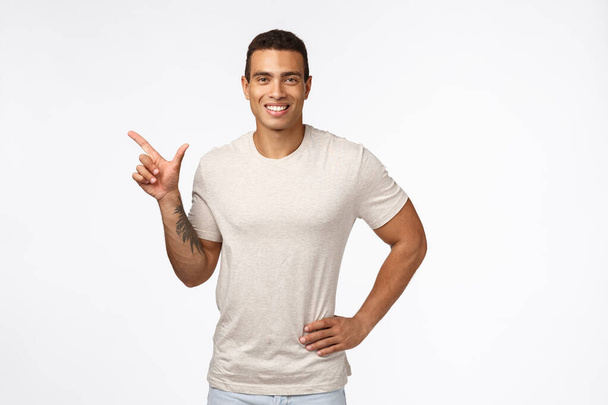 Deportes, personas sanas y concepto movember. Guapo chico hispano sonriente en camiseta casual, tomados de la mano en la cintura relajado, apuntando a la izquierda y sonriendo satisfecho, recomendar promoción, anunciar nuevo producto
 - Foto, Imagen