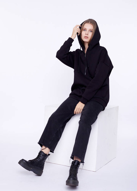 Young girl wearing blank and oversize black long hoody. Sitting on white cube. White background. - Foto, Imagem