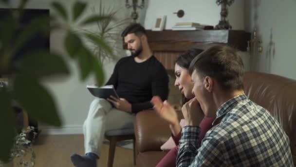 Tired Caucasian family sitting at the foreground and yawning, psychologist taking notes at the background. Young man and woman solving their psychological problems with professional help. - Video
