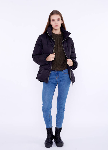 Young girl in a black down jacket and blue jeans isolated on a white background. - 写真・画像