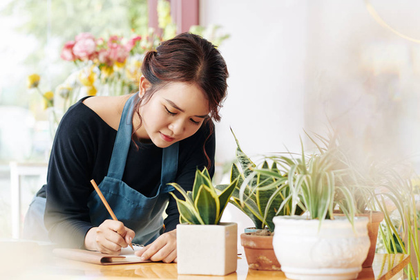 Young Asian flower shop owner writing list of plants and pots to order - Foto, immagini