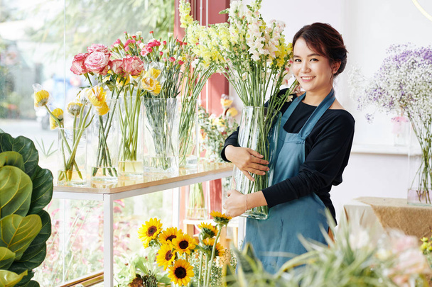Souriant jeune femme asiatique plaçant des vases avec diverses belles fleurs en fleurs dans la boutique de fleurs
 - Photo, image