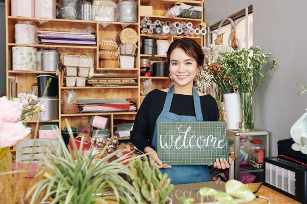 Porträt einer glücklichen jungen Floristin, die an der Theke in einem langsameren Geschäft mit Willkommensschild steht - Foto, Bild