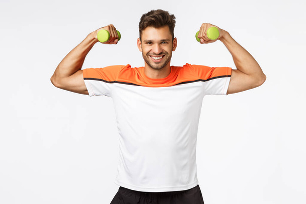 Sport, diet and healthy lifestyle concept. Handsome smiling, happy sportsman enjoy workout, proudly showing his muscles, lift arms and tense biceps to brag perfect body, hold dumbbells at training - 写真・画像