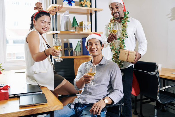 小さなオフィスでクリスマスパーティーを楽しむときにサンタの帽子とトナカイの釣り人を身に着けている多民族ビジネスチーム - 写真・画像