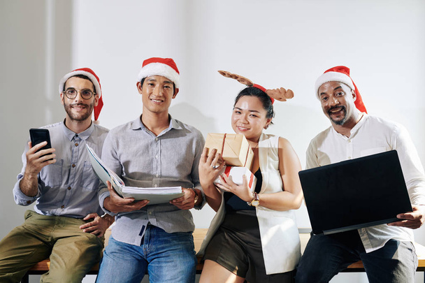 Positives multiethnisches Business-Team in Weihnachtsmützen posiert mit Smartphone, Computer, Dokumentenmappe und Weihnachtsgeschenken - Foto, Bild