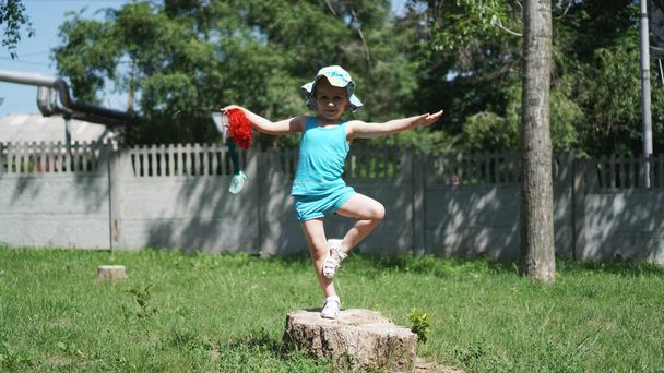ein kleines Mädchen steht auf einem Bein auf einem Baumstumpf in einem Park mit einer Puppe in der Hand - Foto, Bild