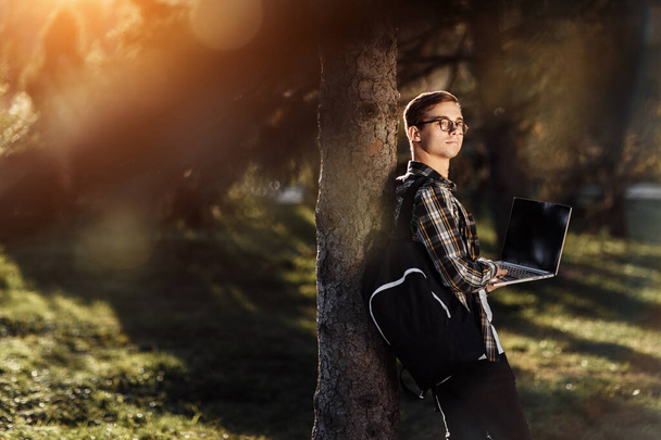 Όμορφος νεαρός φοιτητής σε περιστασιακή στολή κοιτάξουμε μακριά, ενώ στέκεται μπροστά από το δέντρο με σημειωματάριο ή φορητό υπολογιστή στο πάρκο. - Φωτογραφία, εικόνα