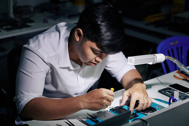 Soustředěný mladý vietnamský opravář pomocí šroubováku sestavit smartphone po jeho opravě - Fotografie, Obrázek