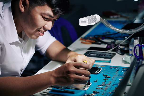 Hymyilevä aasialainen mies korjaa älypuhelinta led-lampun valossa pöydällään
 - Valokuva, kuva