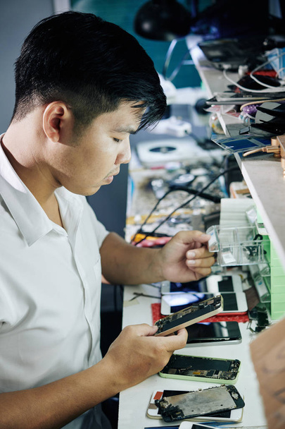 Young Asian repairman opening small drawer when searching for details for broken smartphone - 写真・画像