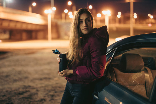 Portrait of woman driver holding thermos bottle leaning on car, night time - Photo, image