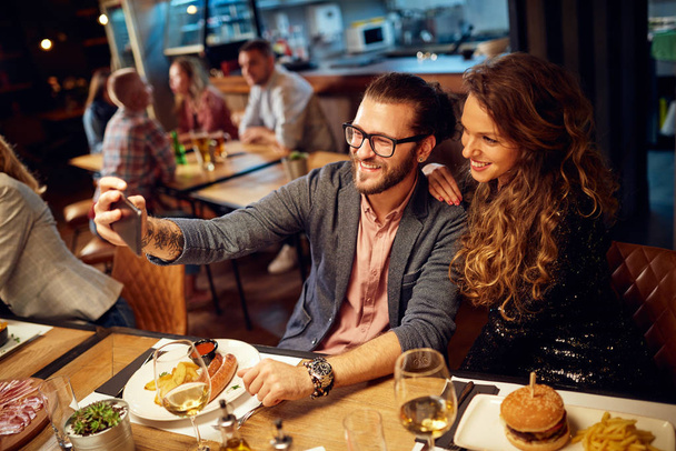 Linda pareja caucásica de moda sentada en el restaurante en la cena y tomando selfie
. - Foto, imagen