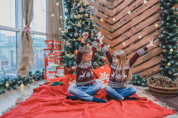 Τέλεια εικόνα όπου το μικρό αγόρι και το κορίτσι γελούν, σηκώνοντας τα χέρια τους σαν νικητές και διασκεδάζοντας μαζί κοντά στο χριστουγεννιάτικο δέντρο. - Φωτογραφία, εικόνα