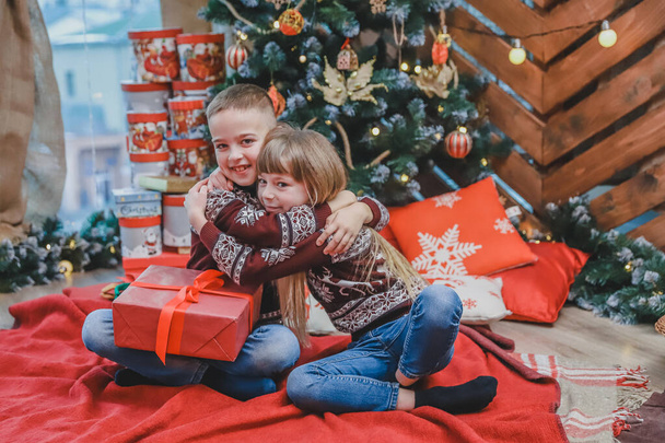 Image of brother and sister in santa hats, tenderly embracing, sharing Christmas presents over wooden background. - Foto, imagen