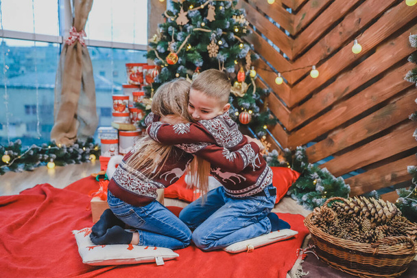 Cute photo of brother and sister hugging at fir-tree on Christmas evening. - Foto, Imagen