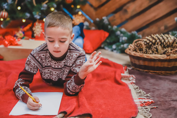 Cute little kid in festive dress lying on the floor, writing letter to Santa. Christmas tree with lights and gifts at the background. - Photo, image